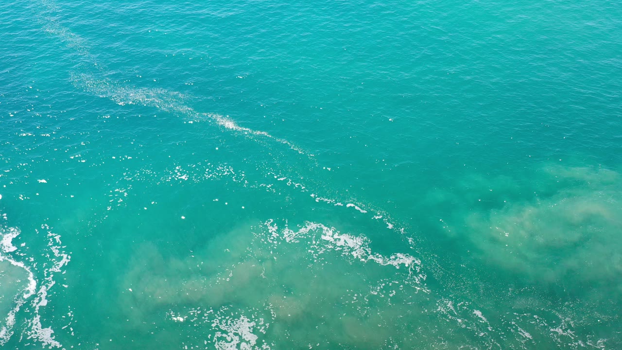 Overhead view of turquoise ocean water in Los Cabos, Mexico, with pale submerged sand patches, scattered white foam, bright surface glints, and no visible people.