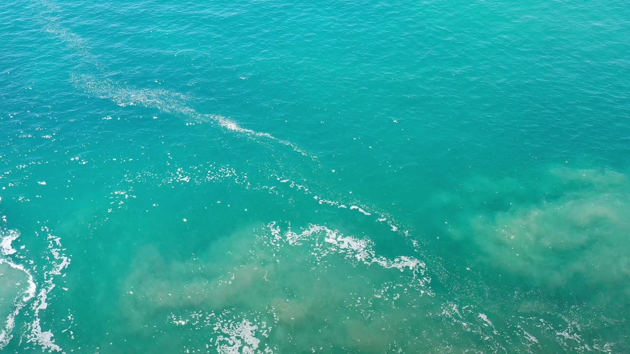 Overhead Los Cabos ocean frame showing open turquoise water, a thin diagonal white foam trace, muted sandy color beneath the surface, and no visible people.