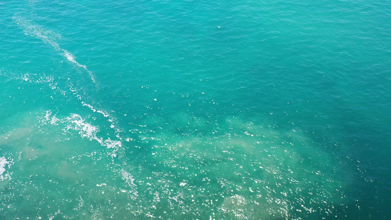 Overhead view of turquoise ocean in Los Cabos, Mexico, with a diagonal white foam streak, pale underwater patches, sun glints, and no visible people.