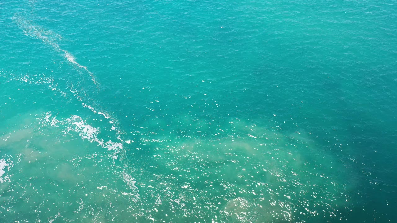 Top-down Los Cabos ocean frame with blue-green water, a broken foam line near the left side, pale sandy shapes below the surface, and no visible people.