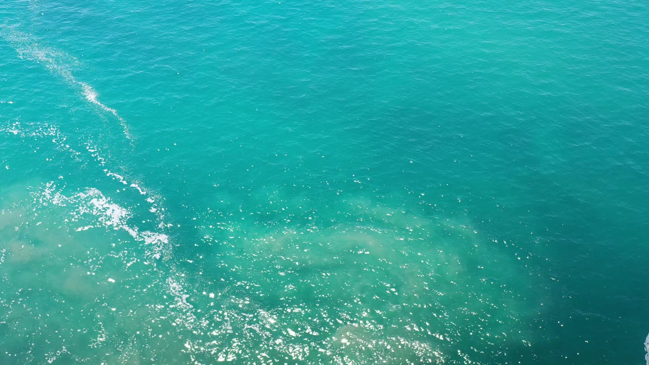 Aerial sea surface with turquoise water, foam streaks, and sunlight.