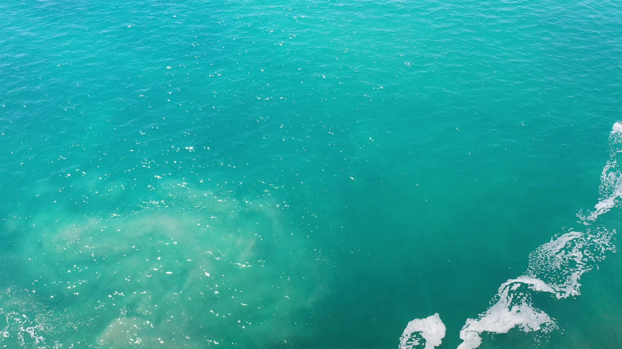 Overhead view of turquoise ocean in Los Cabos, Mexico, with pale underwater sand patches, a small white foam trail near the lower right, and no visible people.