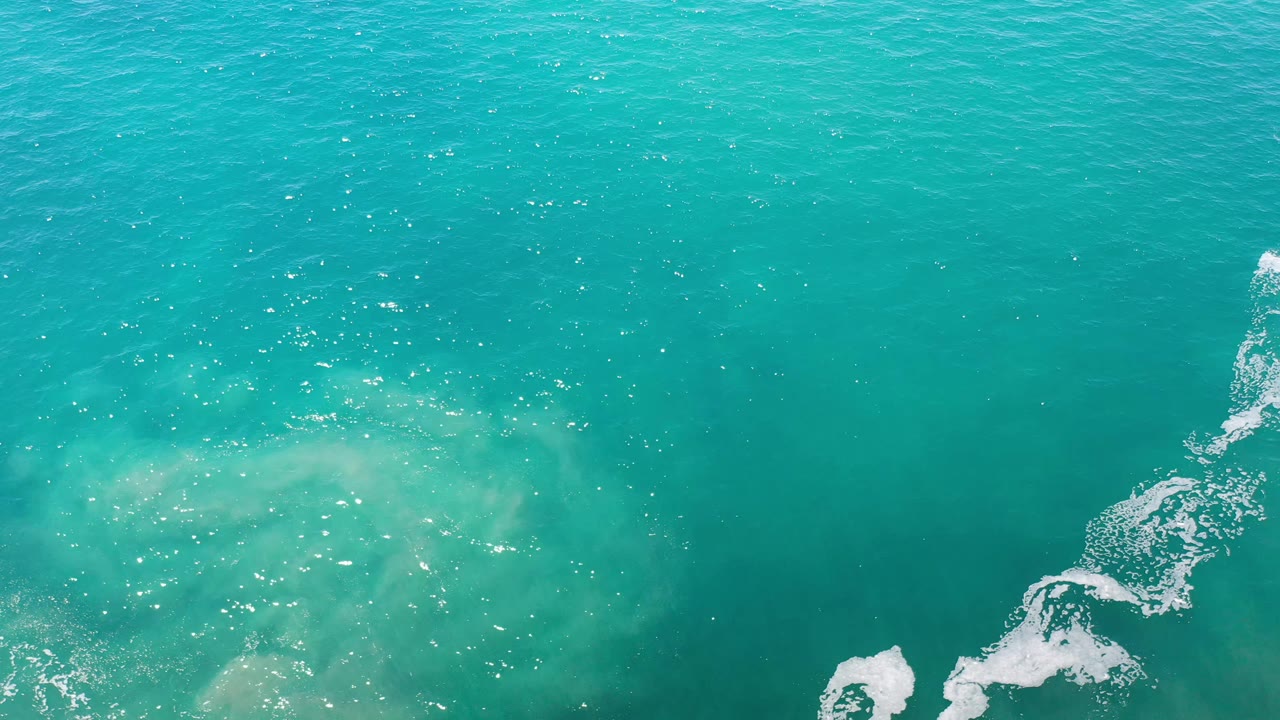 Overhead Los Cabos ocean surface with scattered sun glints, muted sand shapes beneath the water, a thin white foam line toward the right, and no visible people.