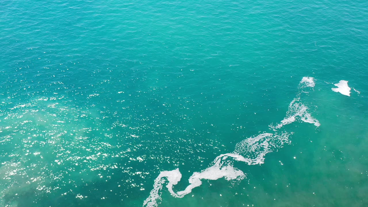 Overhead Los Cabos ocean frame with blue-green water, a broken white foam band near the bottom edge, smaller foam marks near the upper center, and no visible people.