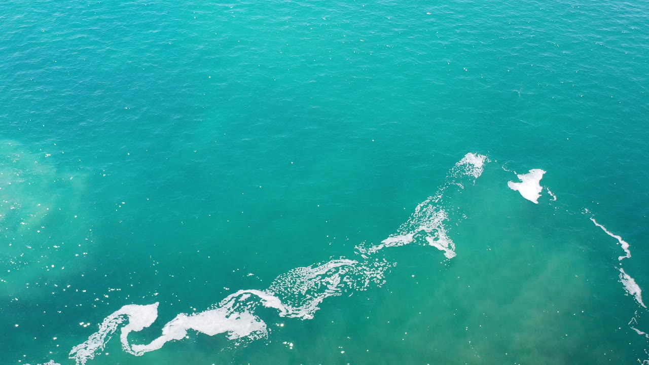 Overhead view of turquoise green ocean water in Los Cabos, Mexico, with white foam trailing near the lower frame and no visible people.