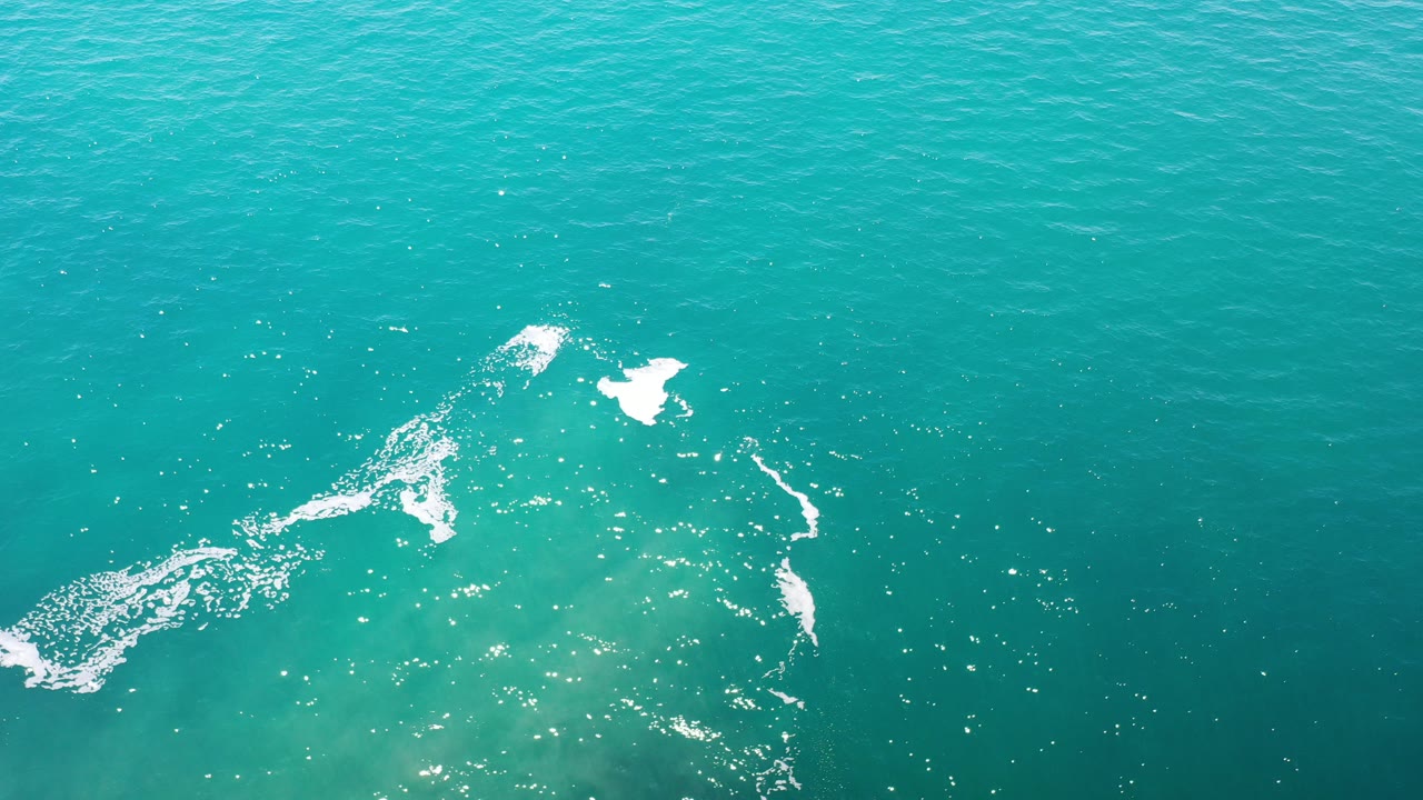 Overhead view of turquoise ocean water in Los Cabos, Mexico, with white foam streaks crossing the lower left and upper center, no visible people.