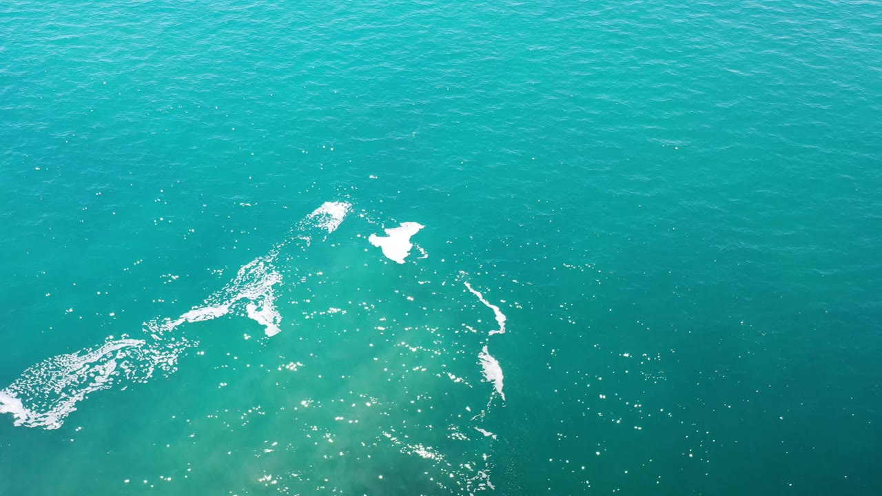 Aerial frame of blue-green ocean in Los Cabos, Mexico, with a loose white foam shape near the upper center and scattered light reflections, no visible people.