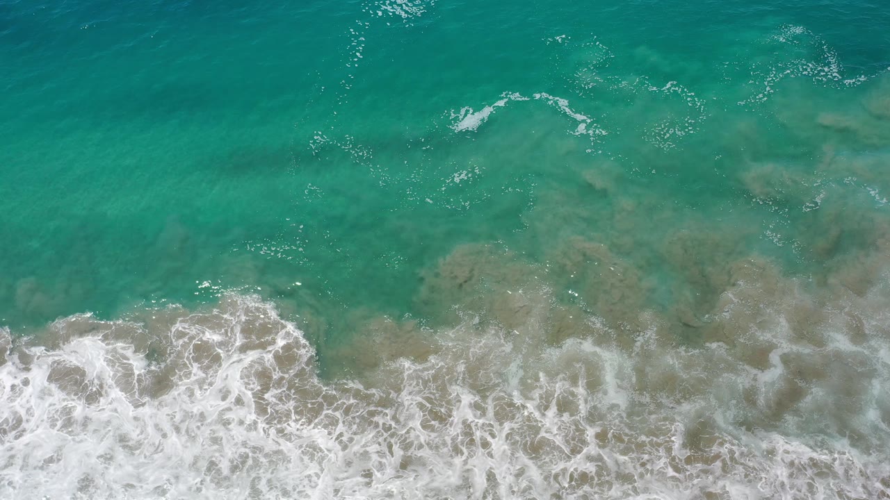 Aerial frame of Los Cabos green water with a thicker band of white foam rolling up from the bottom and no visible people.
