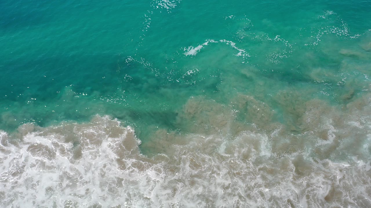 Overhead view of turquoise water and breaking white foam along the Los Cabos coast, with sandy shallows visible below the surface and no visible people.
