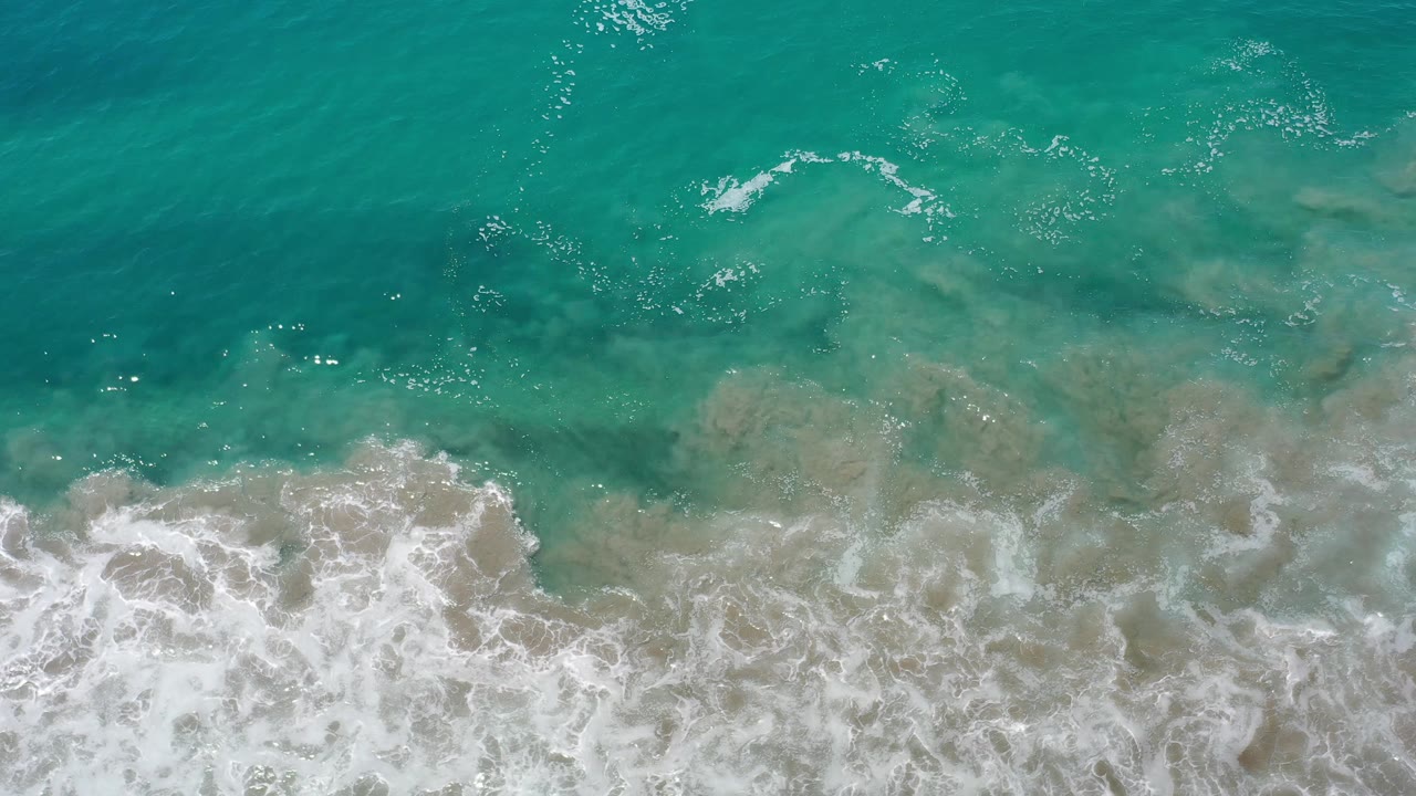 Aerial view over Los Cabos water where foamy surf spreads across the lower frame beneath clearer teal water, with no visible people.