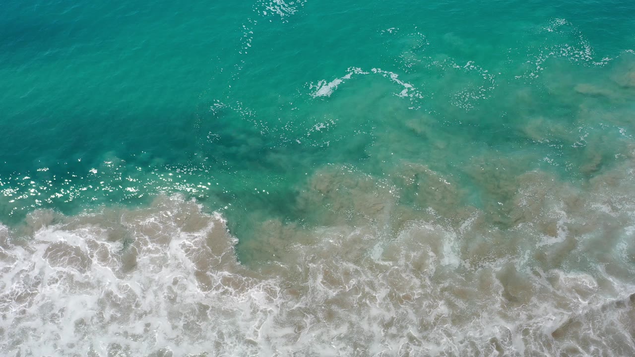 Overhead coastal water with white wave foam and pale sand visible below.
