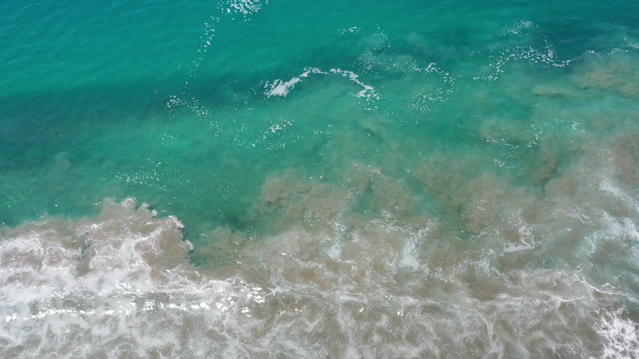 Overhead view of green-blue ocean water in Los Cabos, Mexico, with white foam breaking over tan shallows and no visible people.