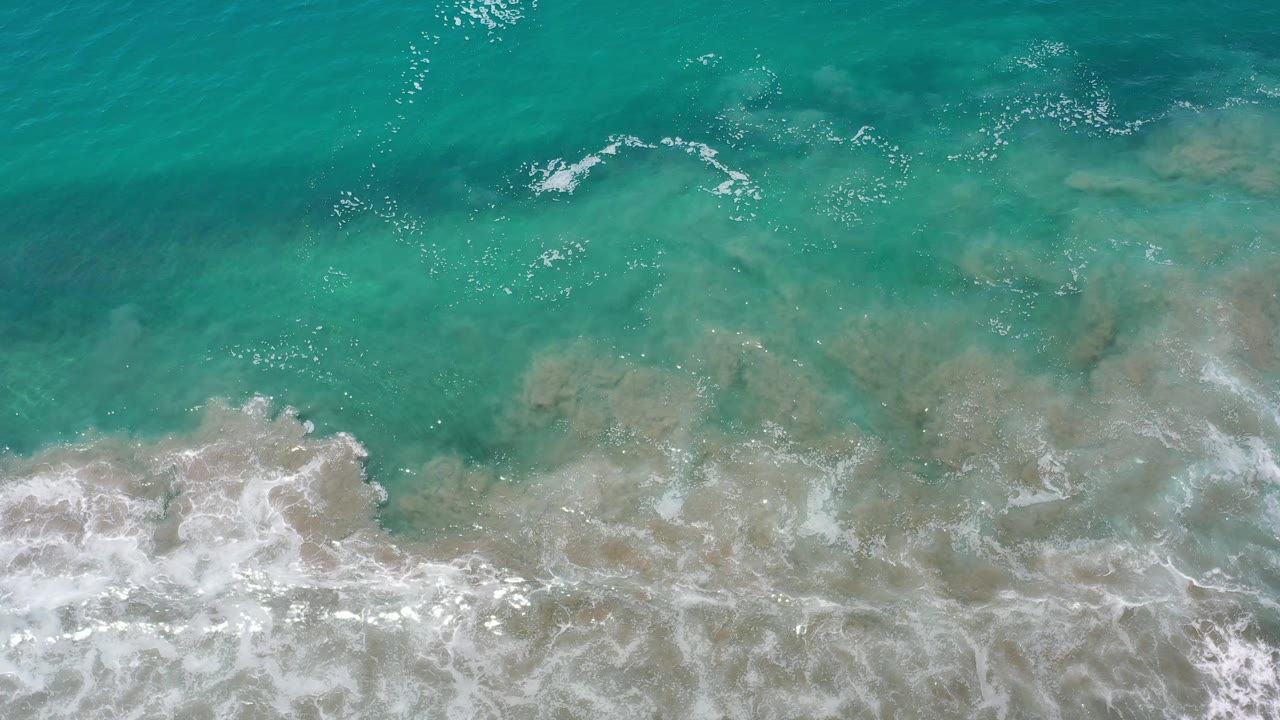 Aerial Los Cabos surf frame with a wide white foam band pushing across green water and pale submerged sand, with no visible people.