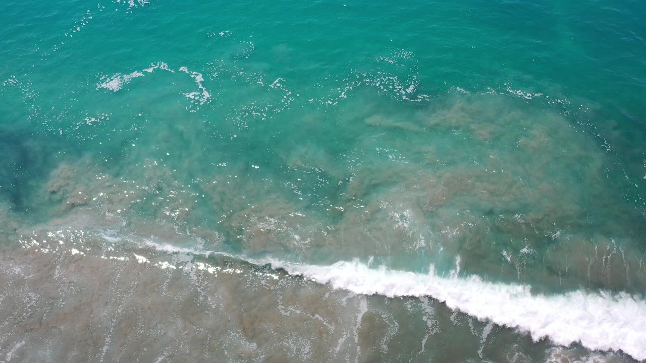 Overhead view of turquoise surf over sandy shallows in Los Cabos, Mexico, with a white wave line entering the lower frame and no visible people.