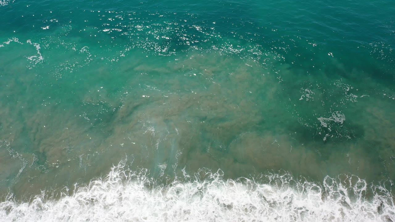 Overhead view of teal ocean water in Los Cabos, Mexico, with pale sandy shallows under the surface, white surf foam along the bottom edge, and no visible people.