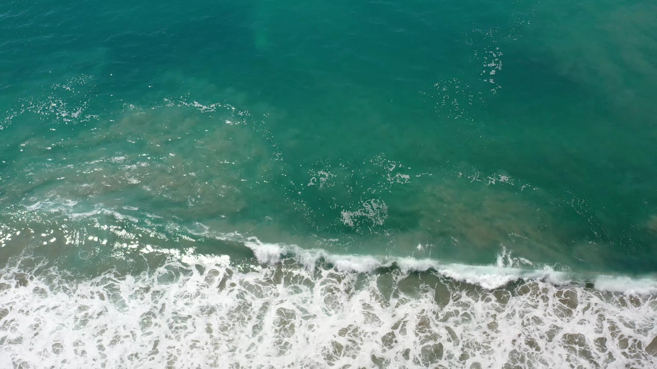 Aerial Los Cabos ocean frame with translucent green water, pale sand visible below, and white-capped surf widening along the bottom with no visible people.