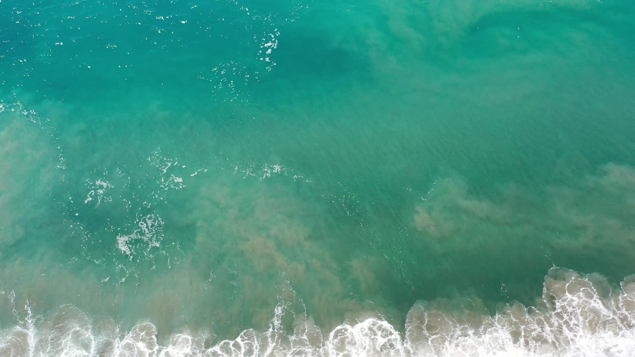 Top-down view of turquoise ocean water in Los Cabos, Mexico, with pale sandy patches below the surface, white foam along the lower edge, and no visible people.