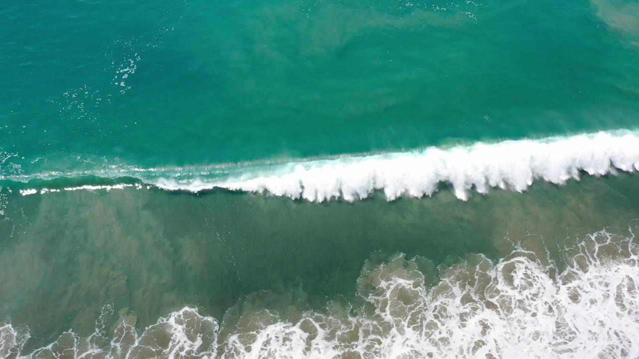 Overhead view of turquoise ocean surf in Los Cabos, Mexico, with a narrow white wave breaking across the frame and no visible people.