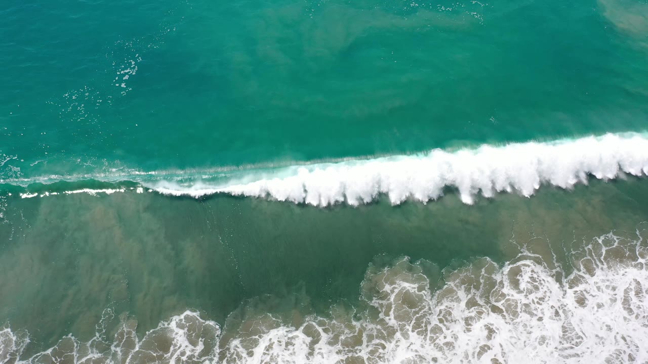 Aerial frame of green-turquoise water in Los Cabos, Mexico, with the white surf line moving across the upper middle and no visible people.
