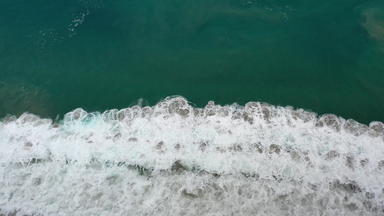 Top-down aerial frame over Los Cabos water, with dark green sea above a horizontal band of foamy white waves and no visible people.