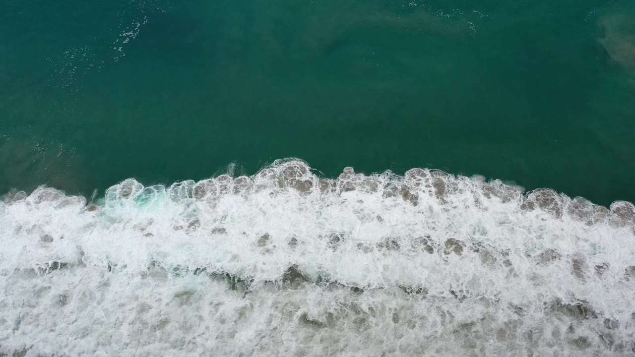 Aerial Los Cabos ocean view with white foam thickening across the middle of the frame, green water above it, and no visible people.
