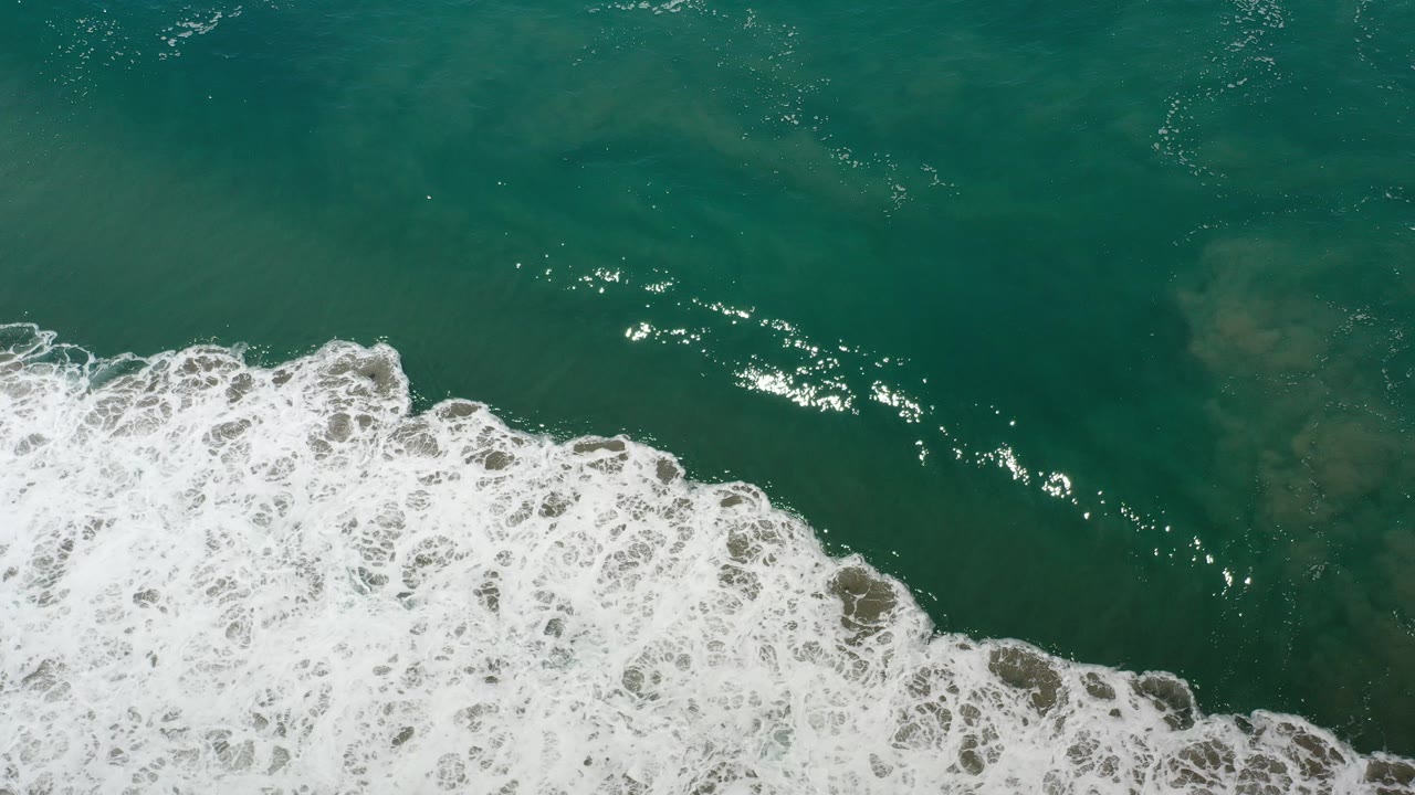 Overhead view of green seawater in Los Cabos, Mexico with white foam breaking across the lower left and no visible people.