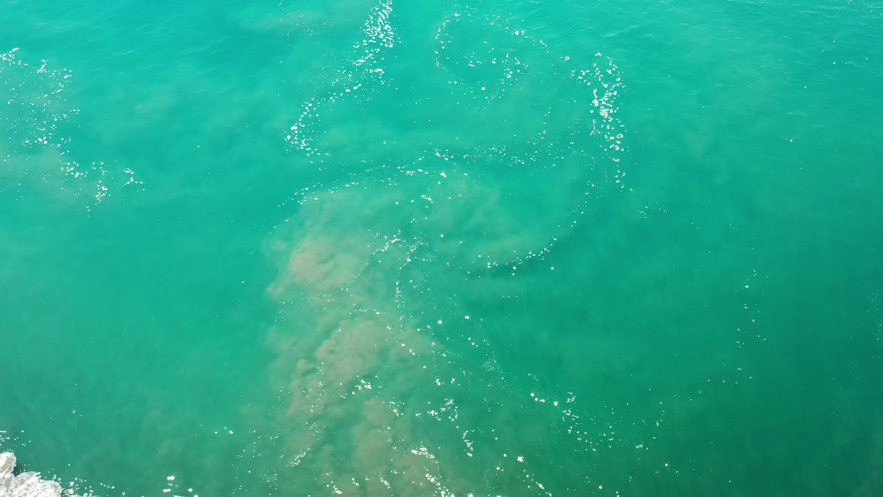 Overhead view of blue-green ocean water in Los Cabos, Mexico, with pale submerged sand patches, scattered white foam, and no visible people.