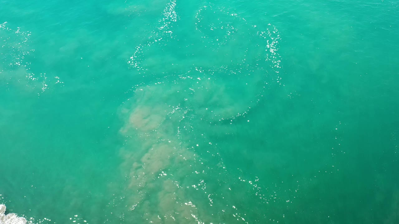 Overhead Los Cabos ocean frame with turquoise surface ripples, bright sun flecks, a pale sand patch below the water, and no visible people.