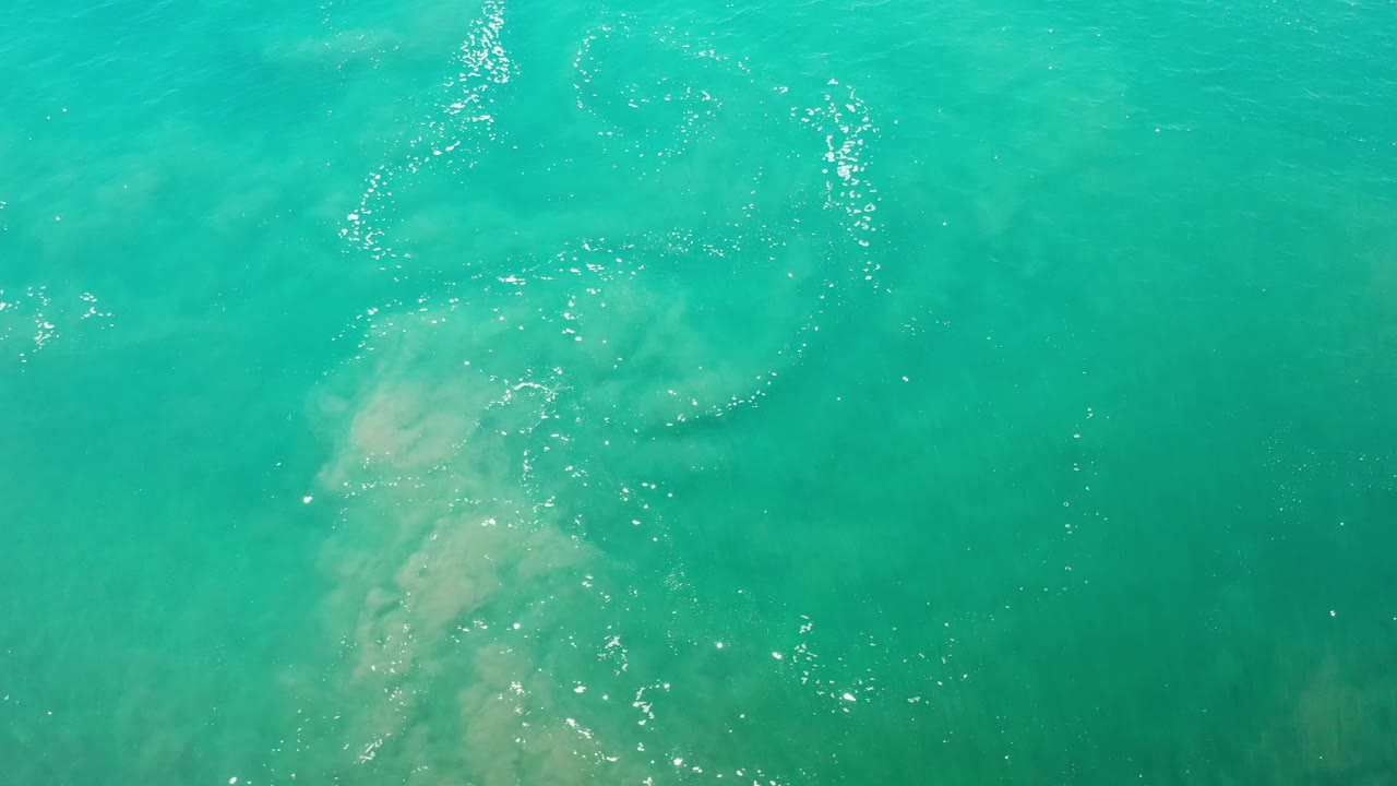 Blue-green Los Cabos water fills the frame, with a curved trail of white foam near the top, cloudy pale sand beneath, and no visible people.