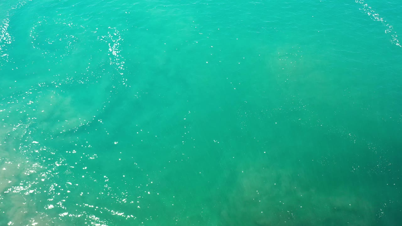 Aerial water surface with bright sparkles and faint pale shapes beneath.