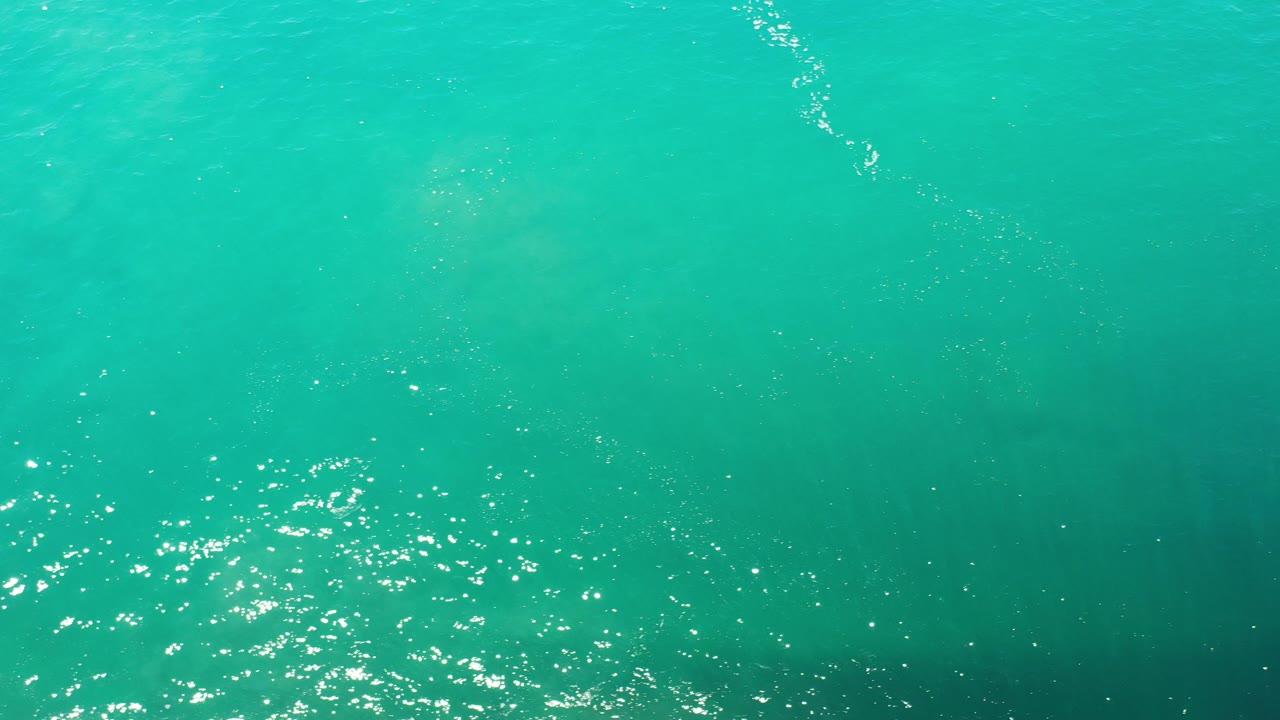 Turquoise green ocean surface with sun sparkle and faint fish below.