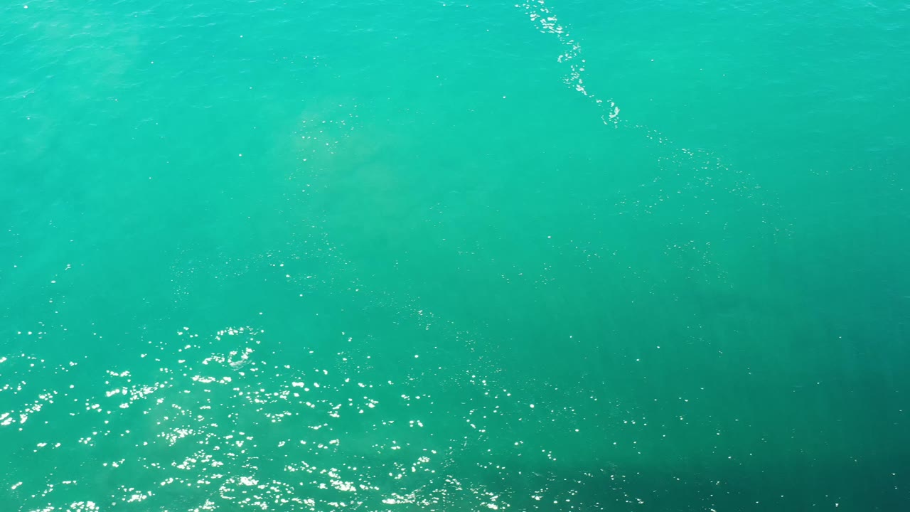 Overhead Los Cabos ocean frame with a wider patch of sparkling ripples near the lower left and faint white fish shapes under green water, no visible people.