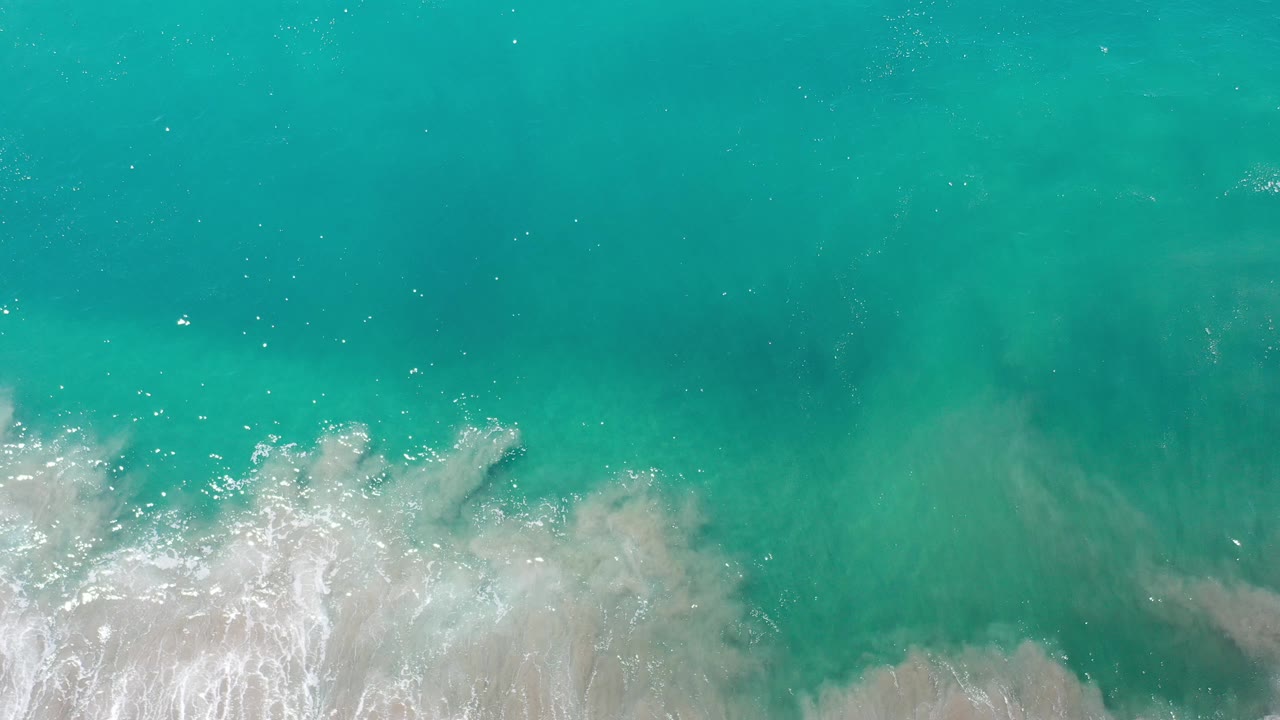 Overhead view of turquoise ocean in Los Cabos, Mexico, with white surf pushing in from the bottom edge and no visible people.