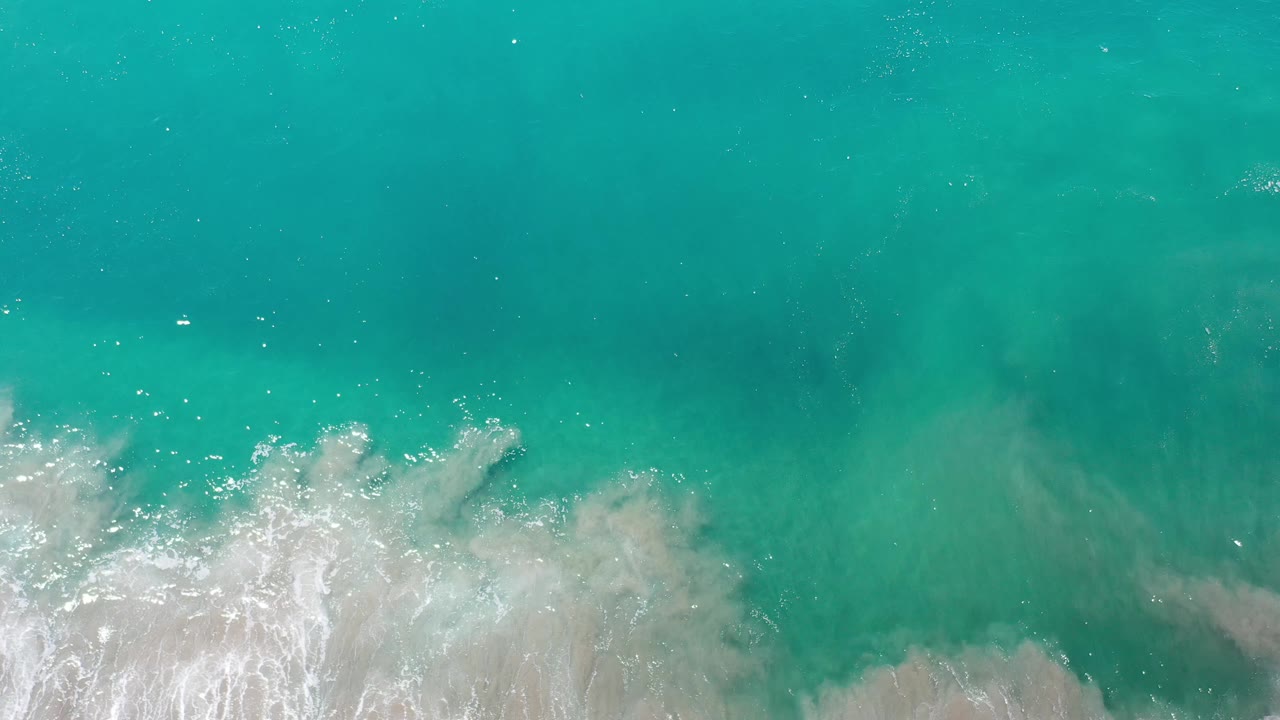 Aerial view of Los Cabos ocean water with foamy white waves spreading wider across the lower frame and no visible people.