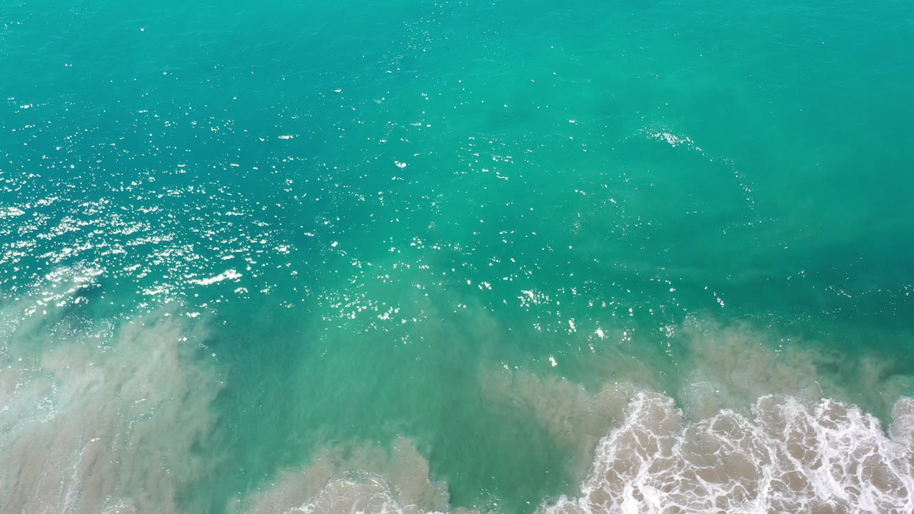 Overhead view of turquoise ocean in Los Cabos, Mexico, with bright sun flecks on the water and white surf beginning to break along the lower edge, no visible people.