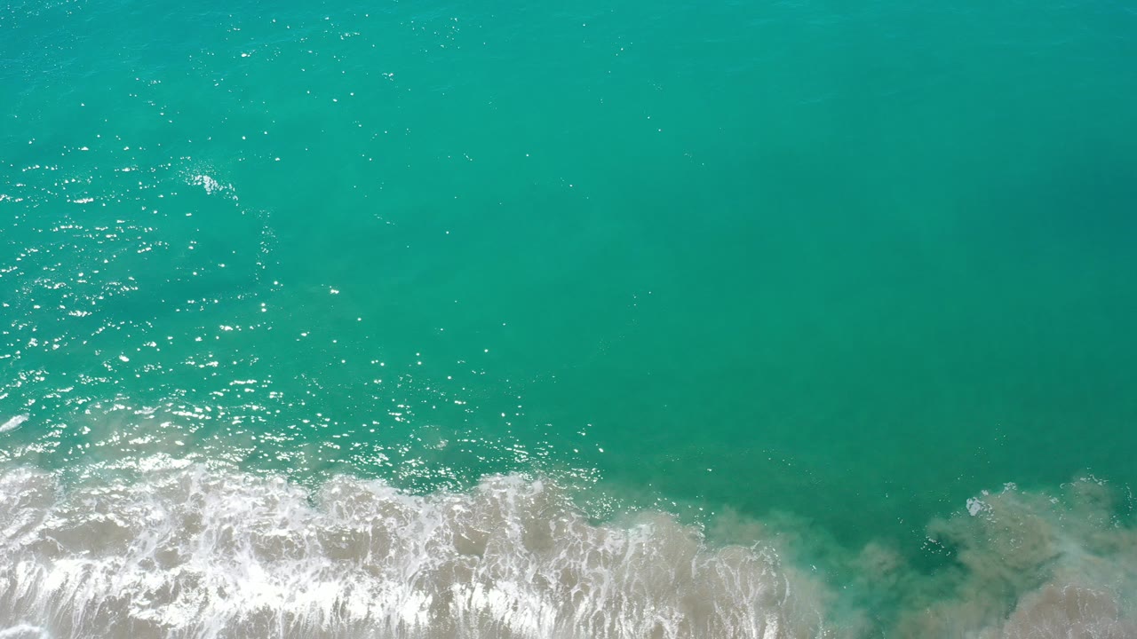 Overhead view of turquoise ocean water in Los Cabos, Mexico, with white surf pushing in from the lower edge and no visible people.
