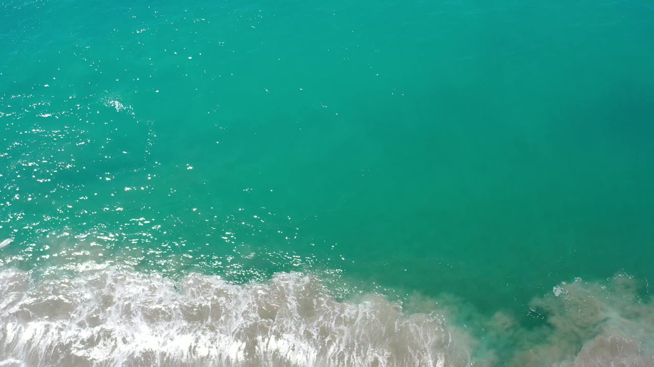 Overhead Los Cabos ocean frame with bright green-blue water, scattered surface sparkle, foamy wave texture near the bottom, and no visible people.