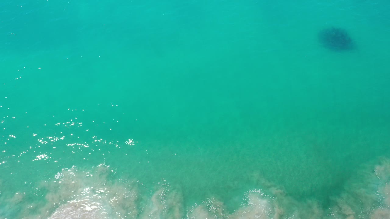 Overhead view of turquoise ocean in Los Cabos, Mexico, with white surf foam entering from the bottom edge and no visible people.