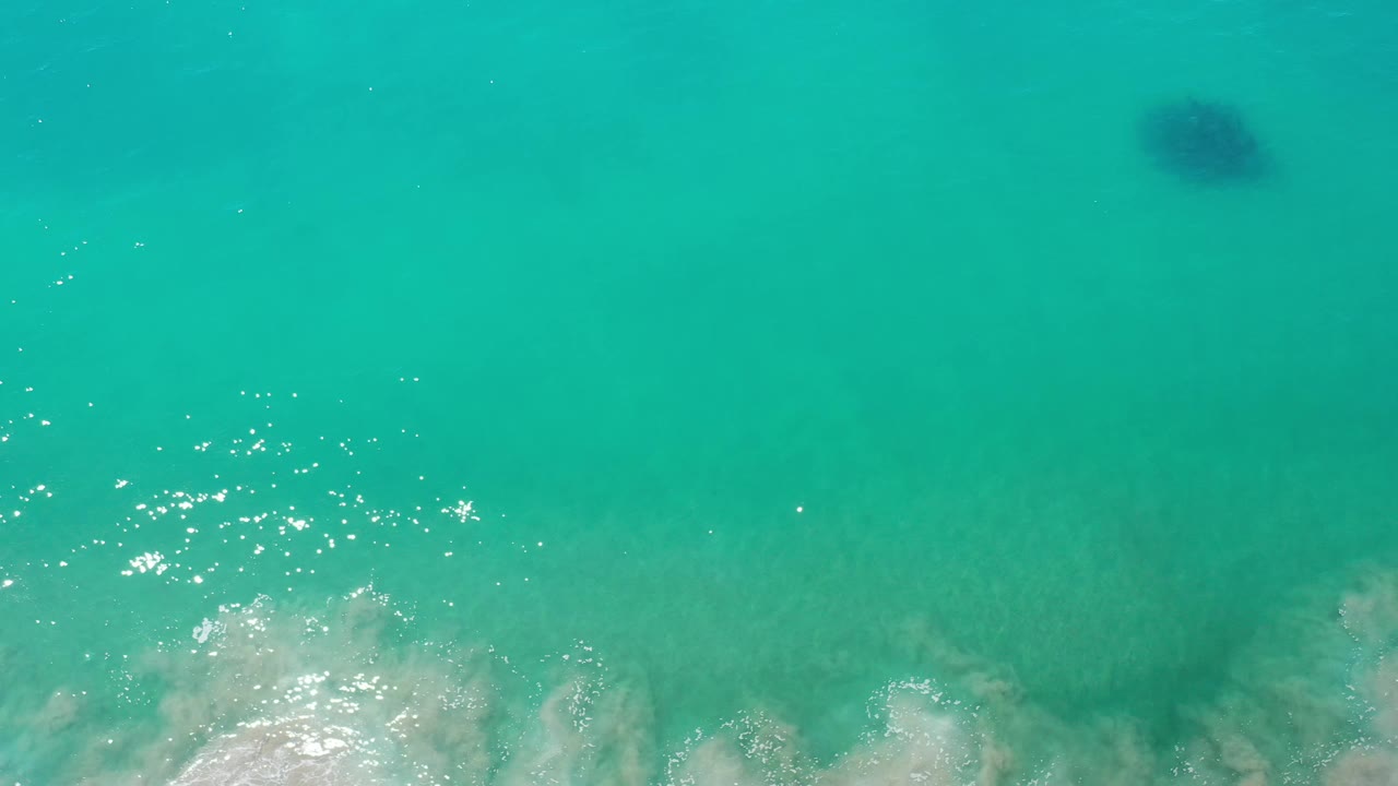 Aerial view of Los Cabos turquoise water with scattered sunlight reflections and pale foam spreading near the lower edge, with no visible people.