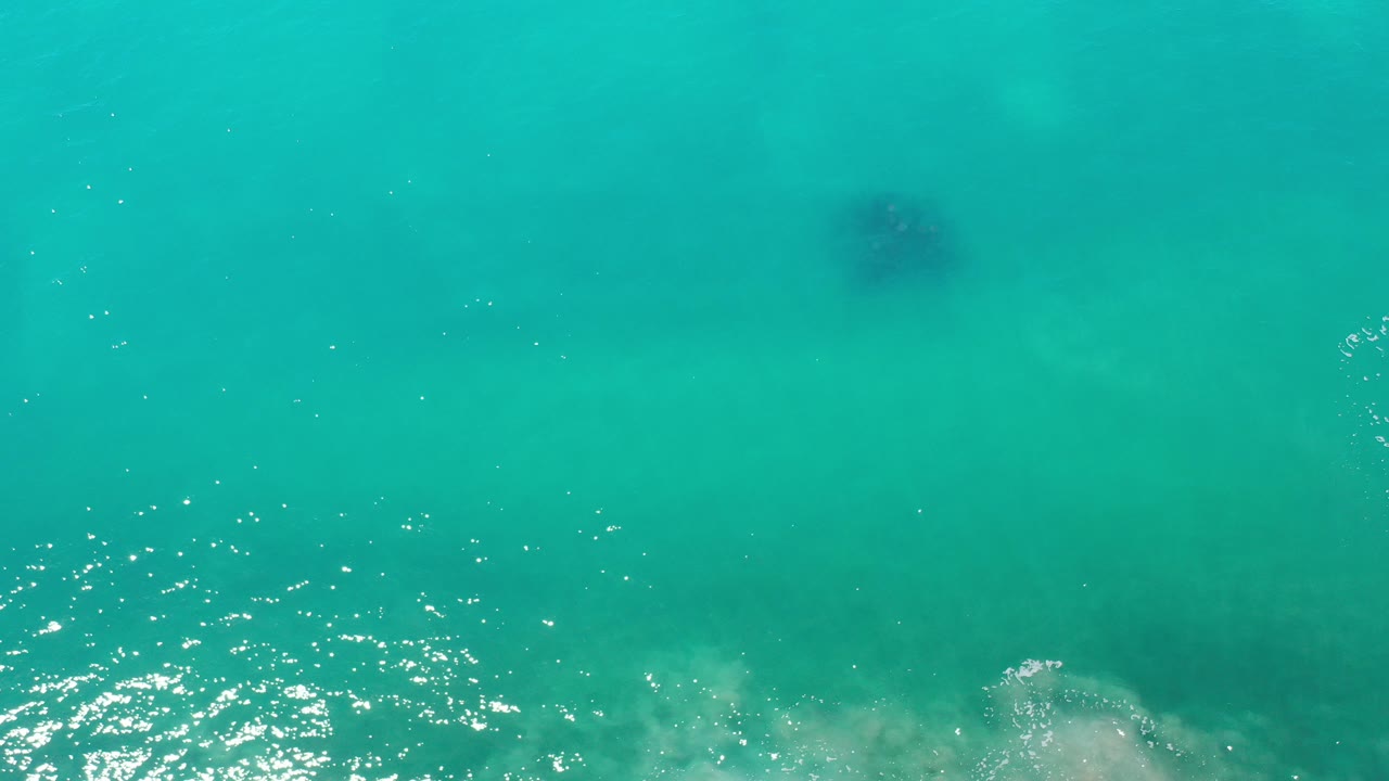 Overhead view of blue-green water in Los Cabos, Mexico, with sun sparkles, faint submerged texture, a dark object near the upper center, and no visible people.