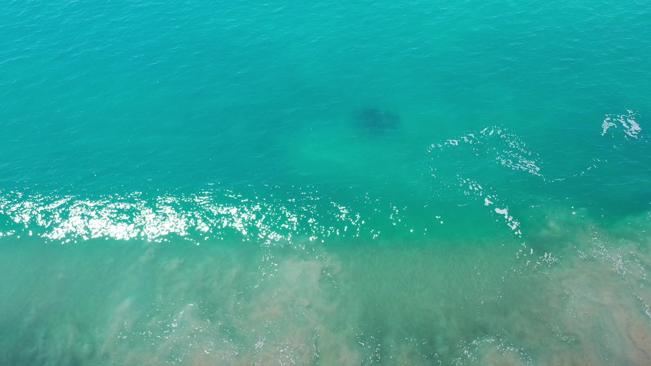 Overhead view of turquoise water in Los Cabos, Mexico, with pale submerged sand near the lower edge, scattered sun sparkle, and no visible people.