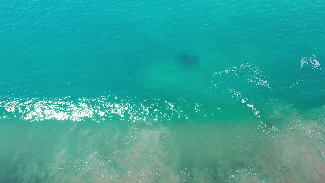 Aerial view over Los Cabos water with turquoise shallows, a darker underwater patch near the upper center, bright surface reflections, and no visible people.