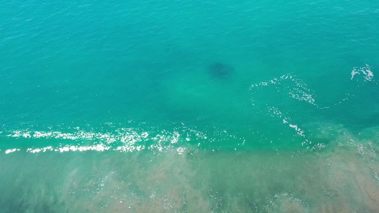 Top-down ocean view with foamy ripples and pale sand visible below the water.