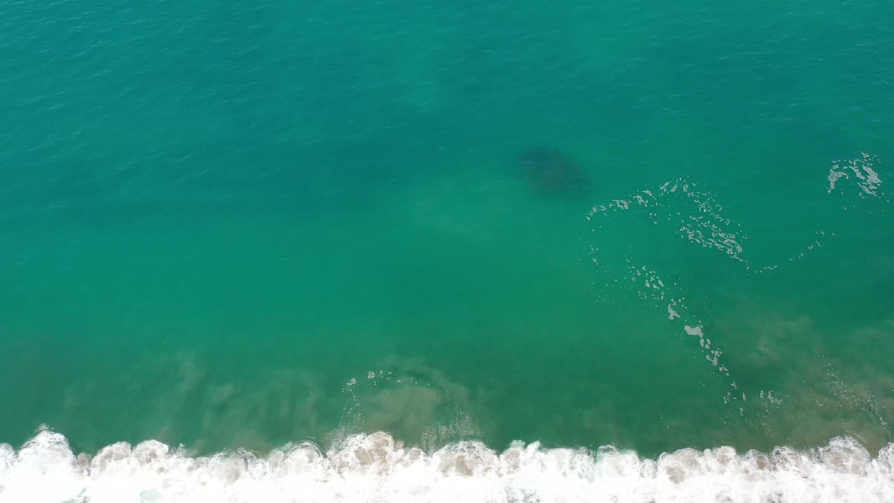 Aerial ocean surface with dark patches and white foam near the shore edge.