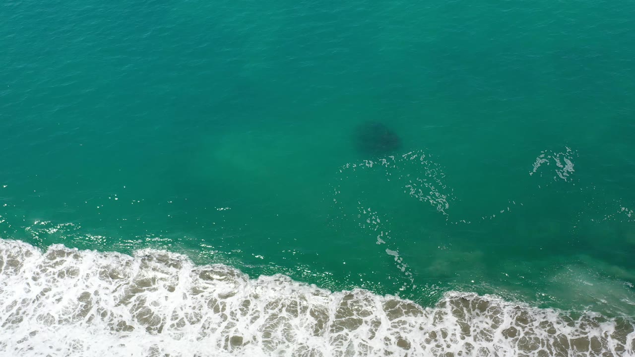 Aerial view over Los Cabos water with foamy surf widening near the bottom and dark underwater patches visible above it, no visible people.