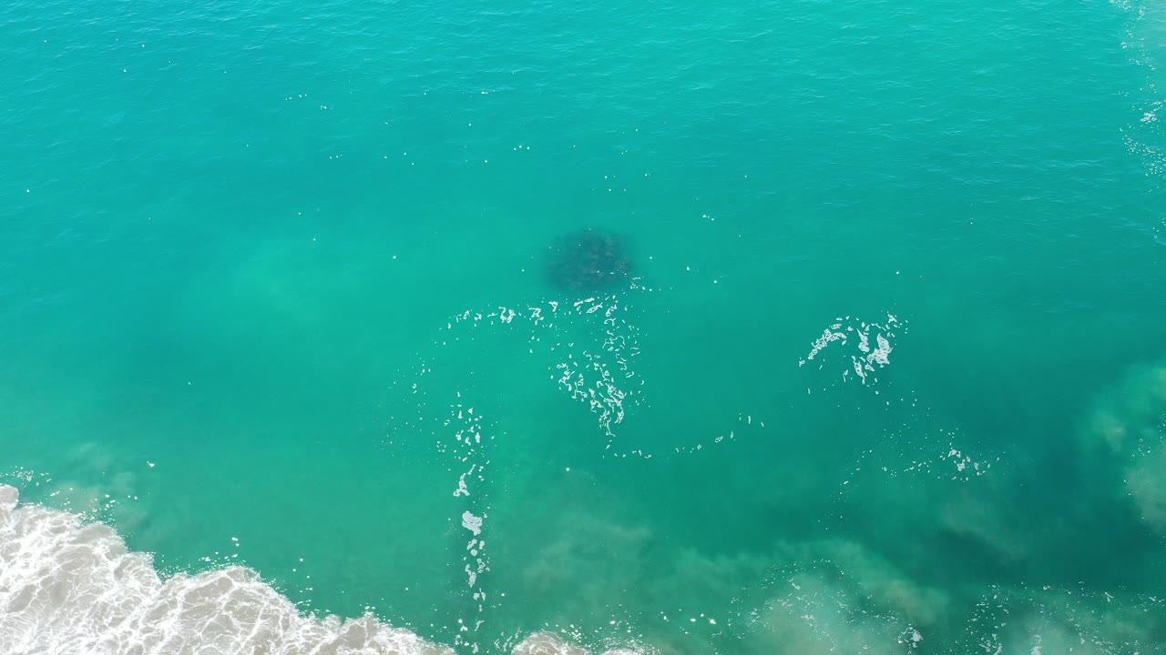 Overhead view of turquoise ocean in Los Cabos, Mexico, with white foam entering from the lower left and dark underwater patches visible, no visible people.