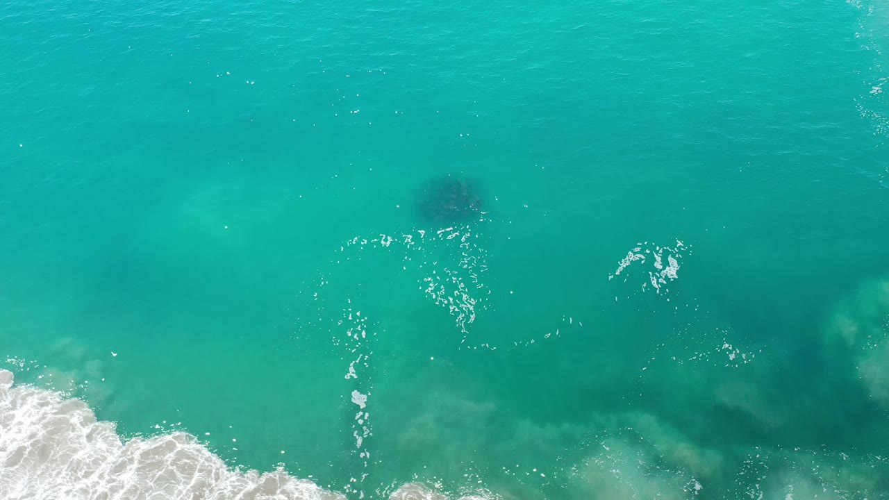 Aerial view of blue-green water in Los Cabos, Mexico, with scattered white foam near the lower left and darker shapes under the surface, no visible people.