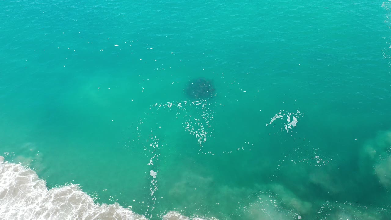 Turquoise ocean surface with bright sun sparkles and surf foam along the lower left.