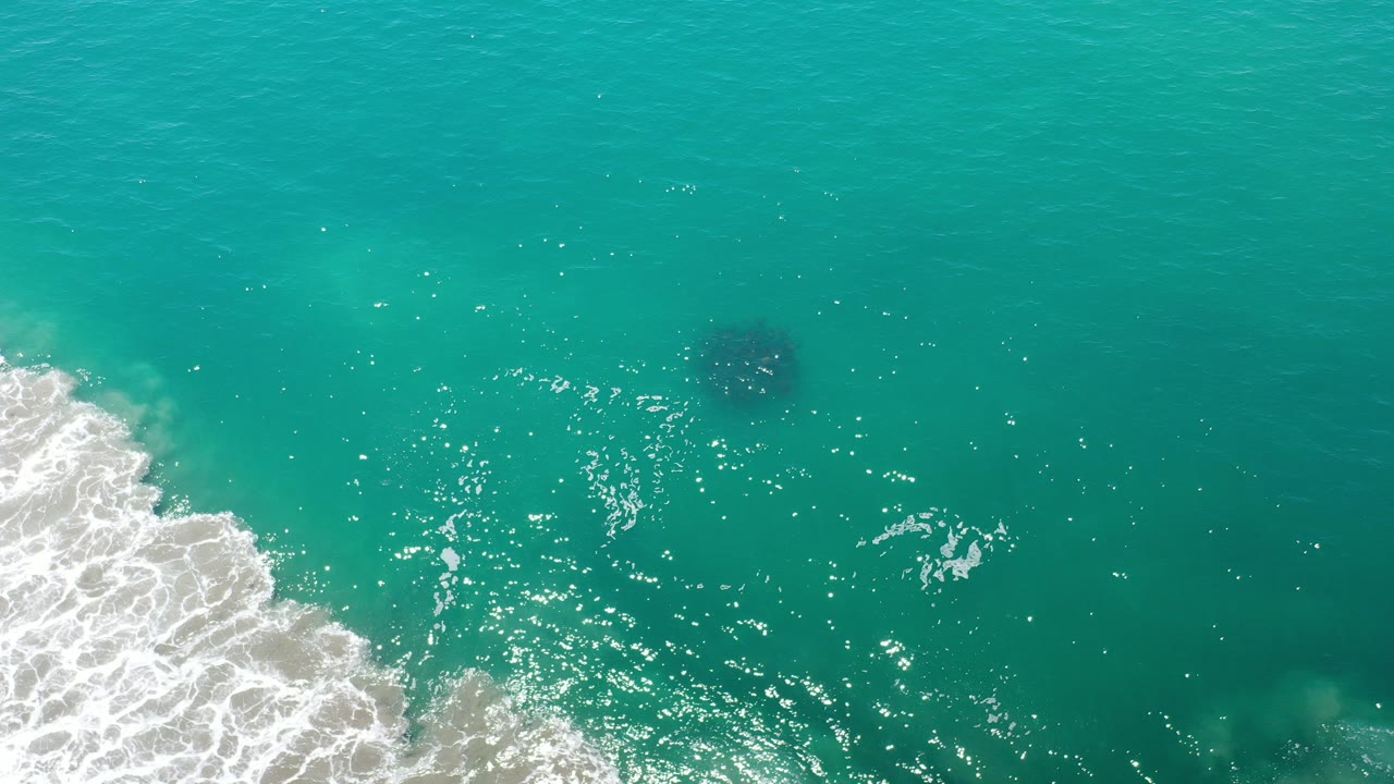 Overhead view of green ocean in Los Cabos, Mexico, with white surf pushing in from the left and a dark circular underwater shape near the upper center, no visible people.