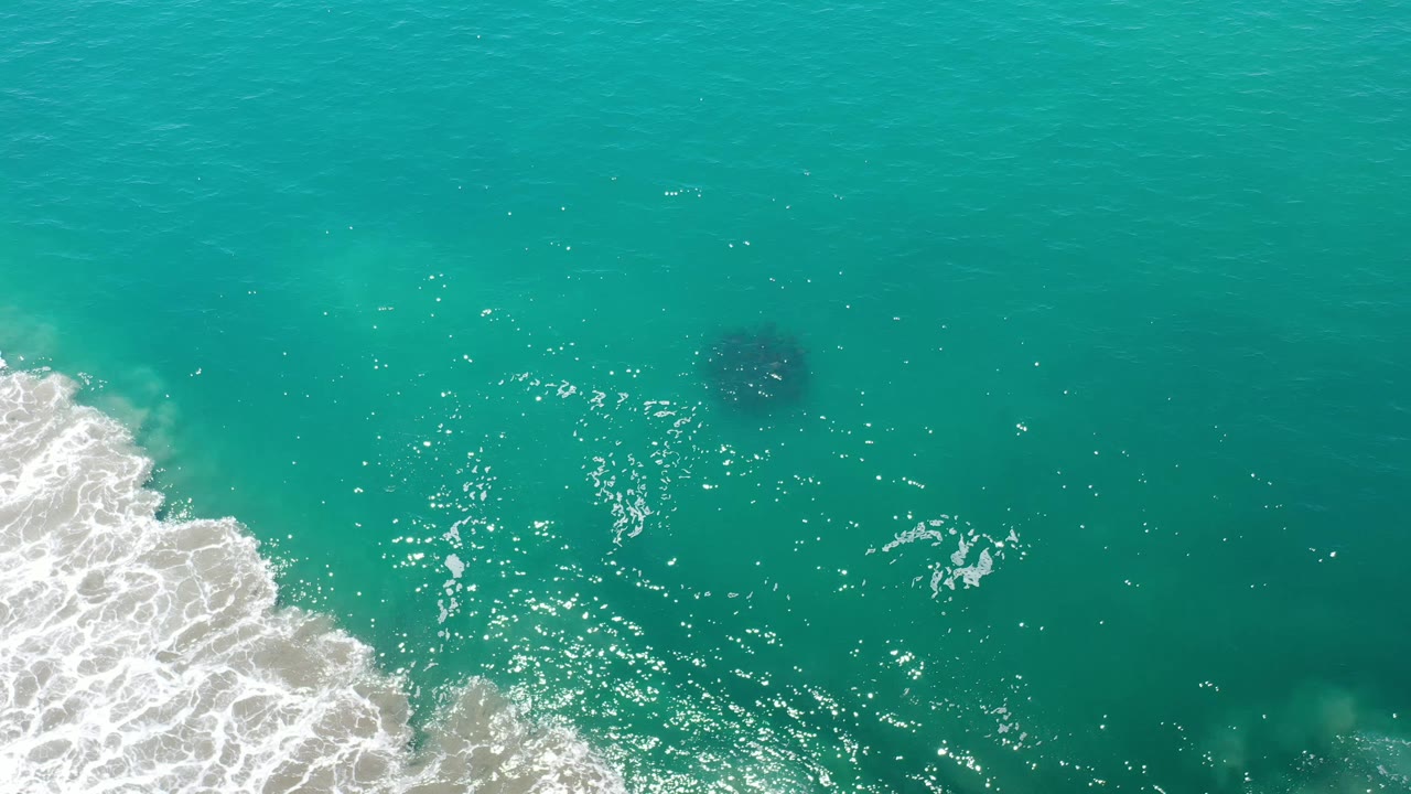 Aerial Los Cabos water view with bright surface reflections, foamy surf along the left side, and the dark underwater form slightly right of center, no visible people.